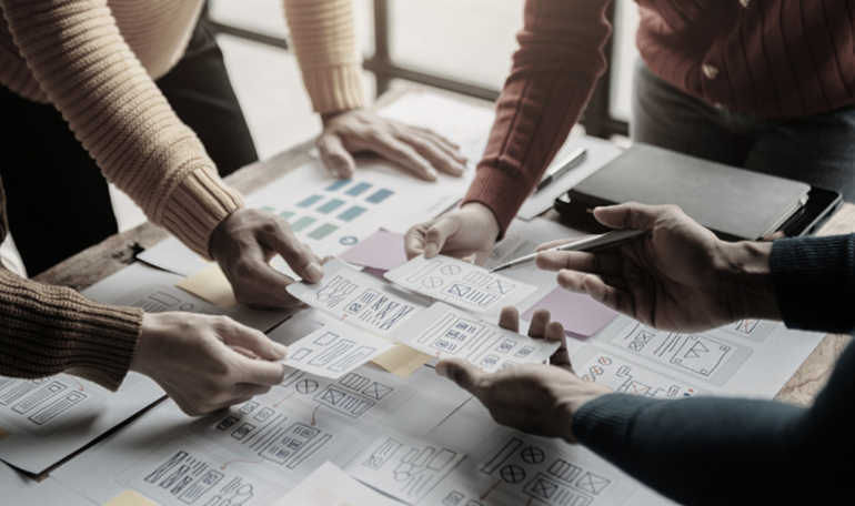 My Digital Solutions – The Global Tech Hub® UK: Team collaboration session with professionals reviewing website and mobile app wireframes, sketches, and layouts on a table, symbolizing creative UI/UX design, digital product development, and innovative design strategy for businesses.